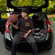 Person sitting in the open trunk of a car holding a basketball, with trees and road in the background.